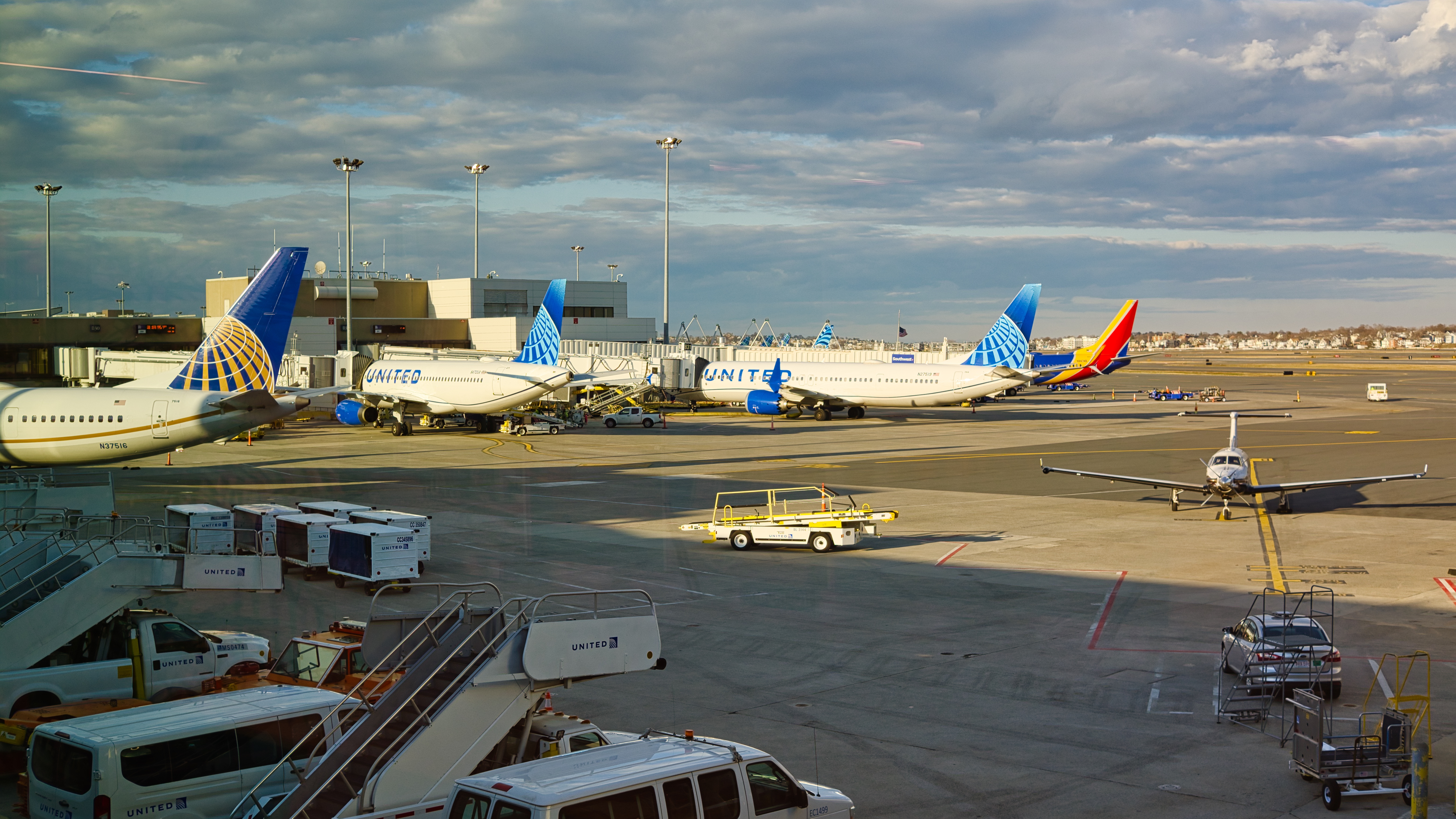 Passenger aircraft parked by Terminal B at Boston Logan, with service vehicles on the apron.