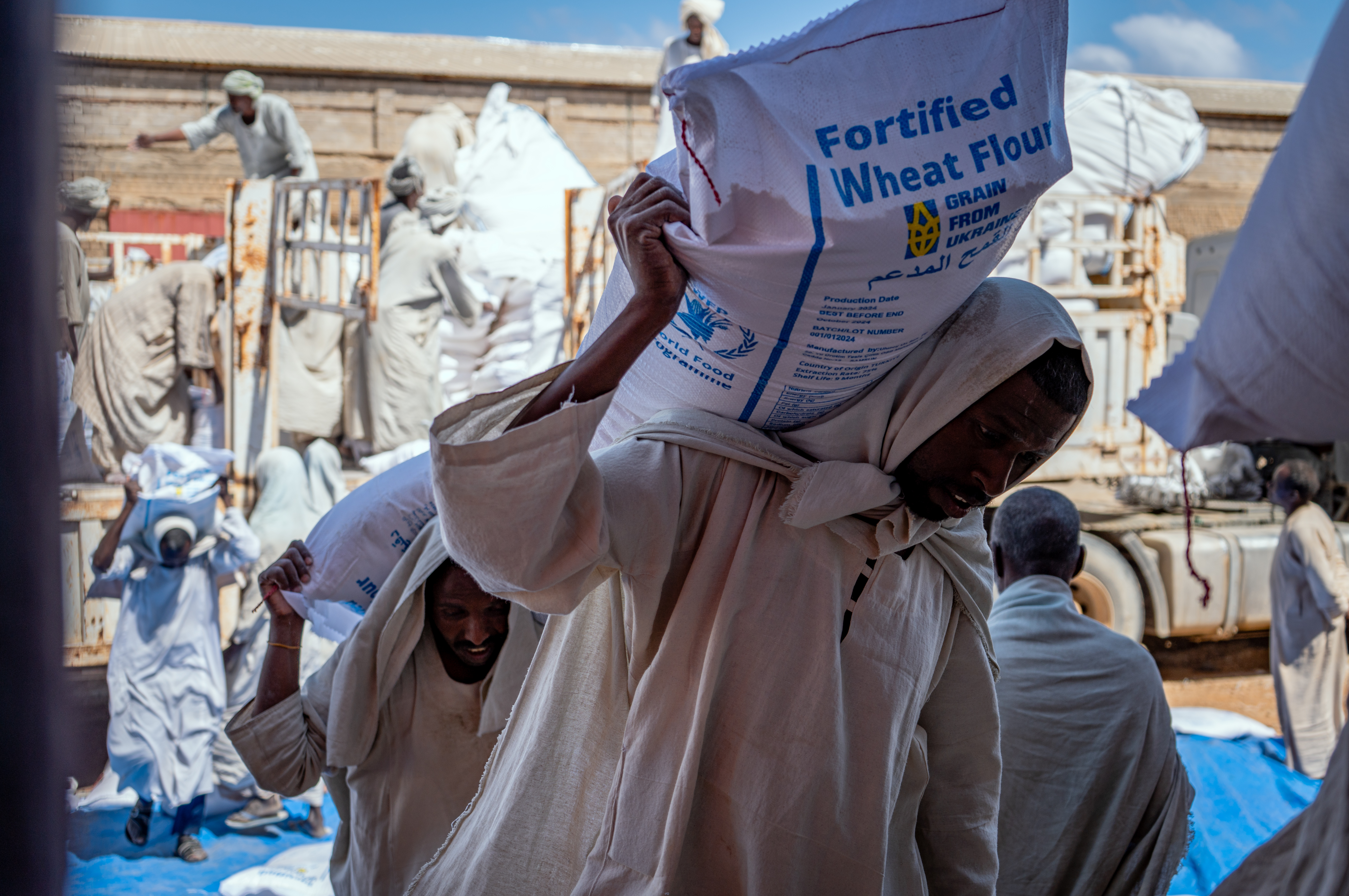 Workers carry sacks of fortified wheat flour near trucks and a warehouse.
