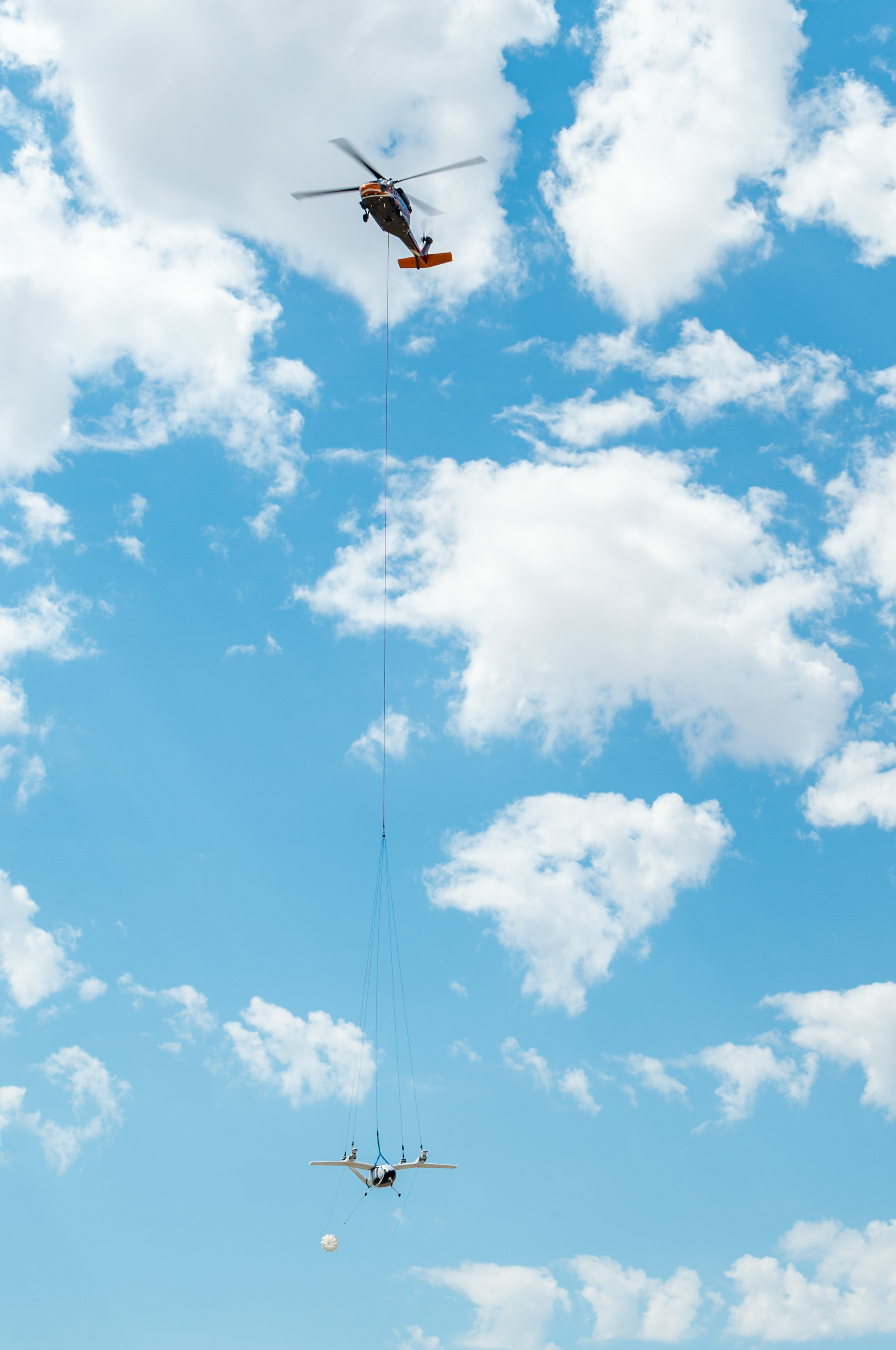 A helicopter carries a small aircraft suspended below long cables against a blue sky.